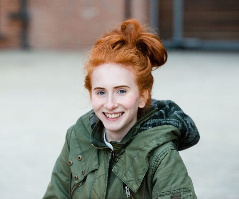 Young woman with vibrant red hair tied up in a bun and wearing a forest green goat is smiling